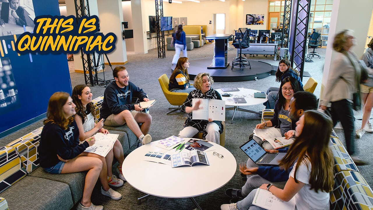 A group of 5 students sitting around a table as a professors shows them something in a book, plays video