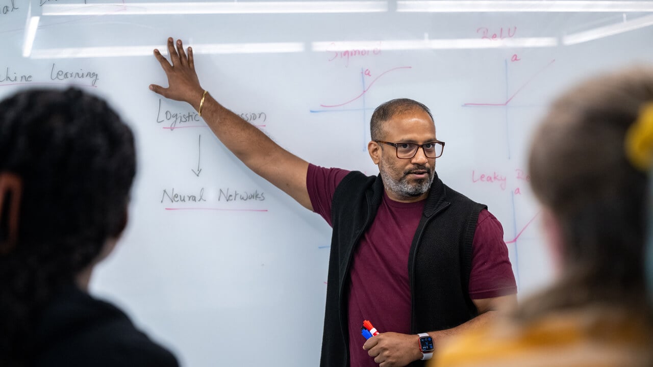 A professor gesturing to a white board in front of a computing class