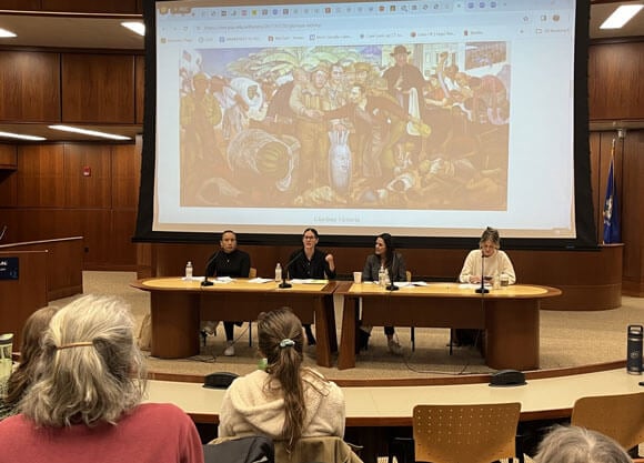 A group of panelists sit in front of a screen at the front of a room.