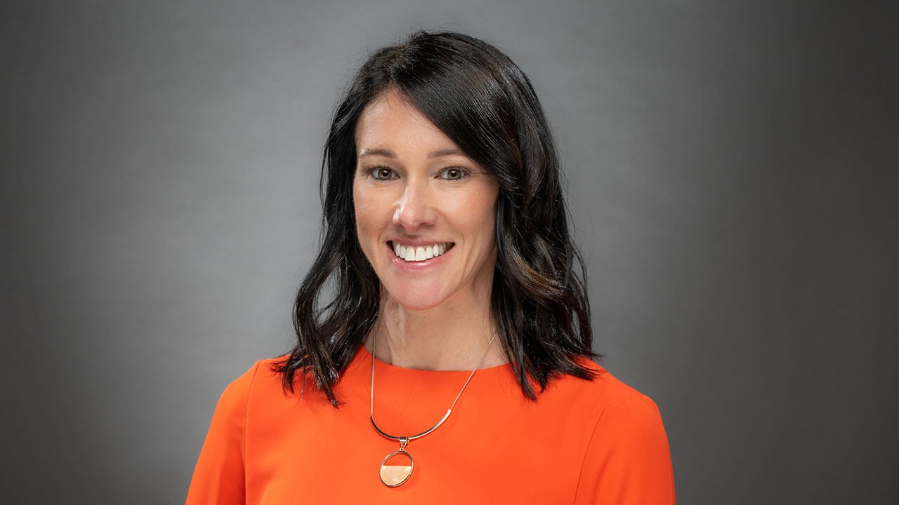 Kori Stewart poses for her picture, smiling, she is a woman with dark brown hair wearing an orange shirt and golden necklace