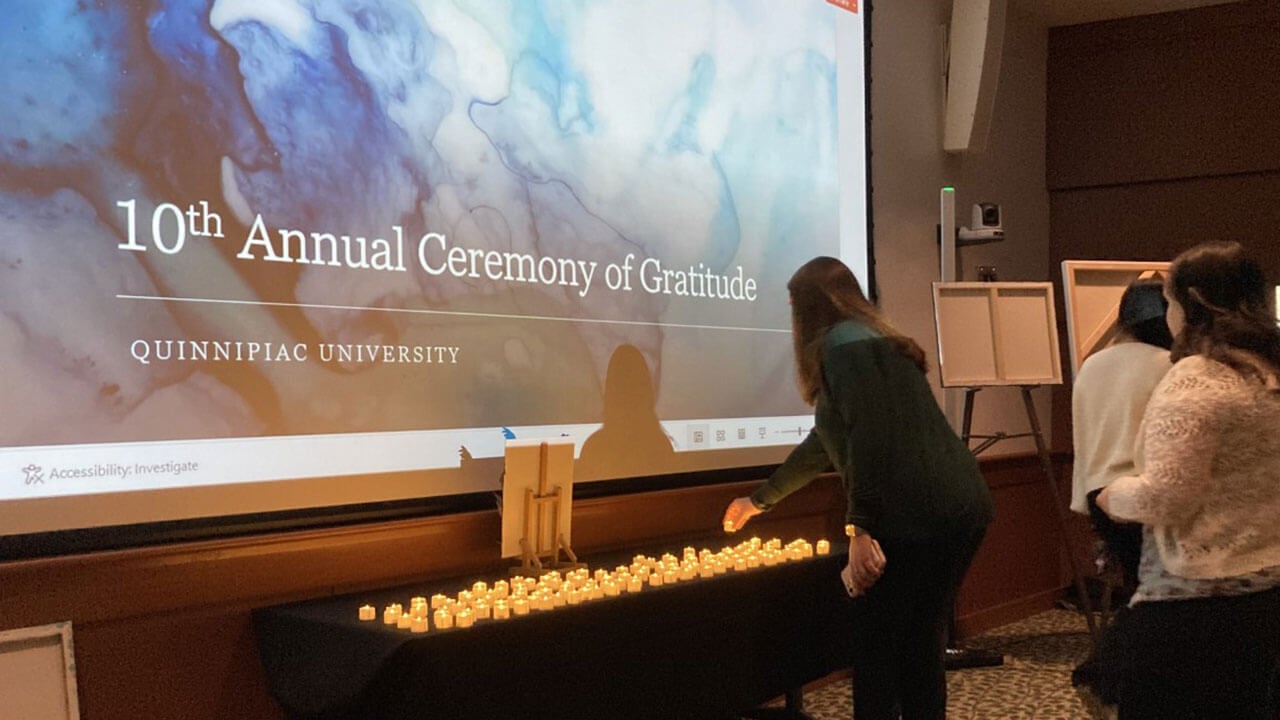 dark haired woman leans down to place lit tealight candle on a table full of them, another woman waits behind her with her own table, projected in front of them is a 10th anniversary watercolor picture