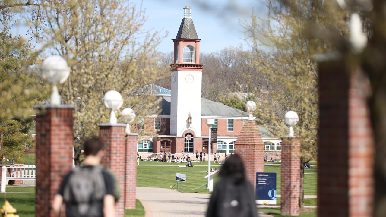 Dozens of students with backpacks cross the Quinnipiac quad on a spring day