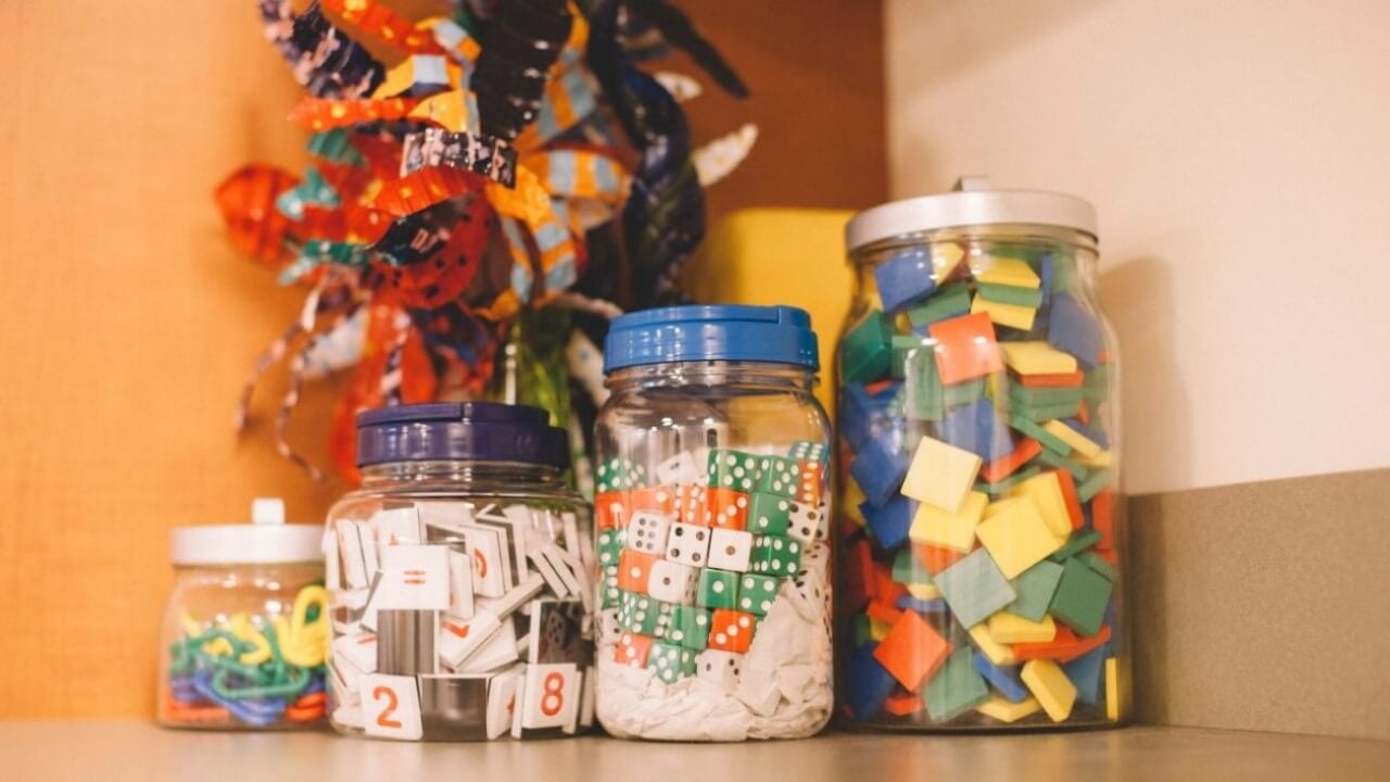 Jars of elementary teaching materials sit on a counter, including dice, large paper clips, and numbered and colored tiles.