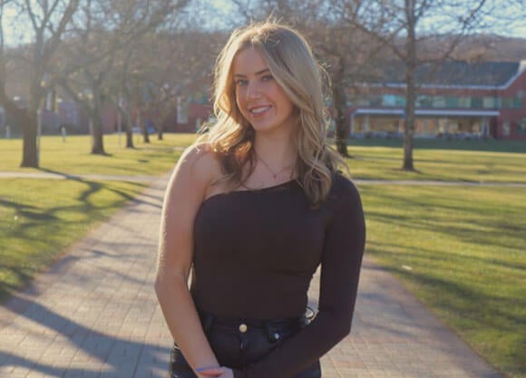 Photo of Quinnipiac student Riley Propersi standing on the mount caramel campus quad with the Echlin Center in the background