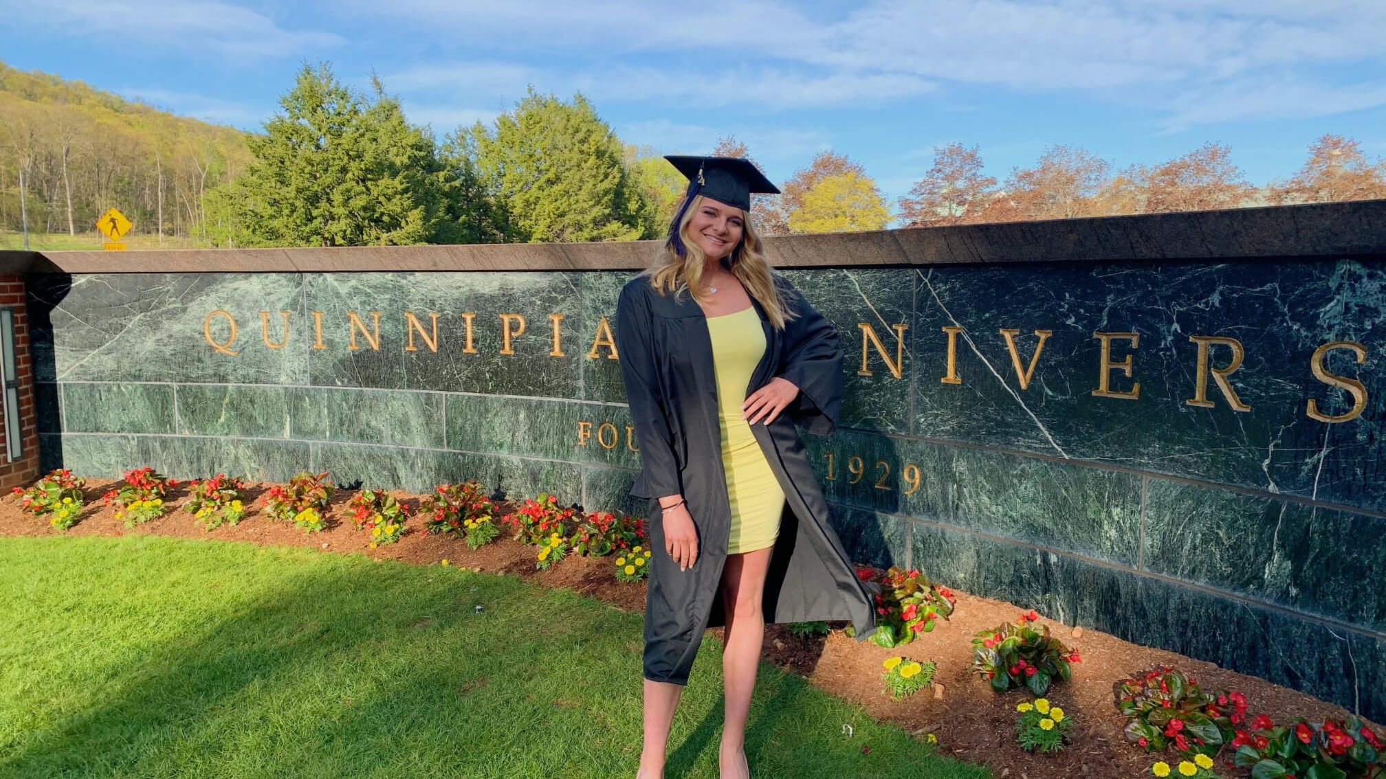 Heather Popovics standing in front of Quinnipiac sign at Mt. Carmel campus in graduation regalia