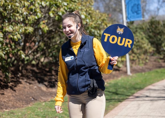 A smiling undergraduate student wears an admissions vest and a headset and holds up a "tour" sign on a sunny day