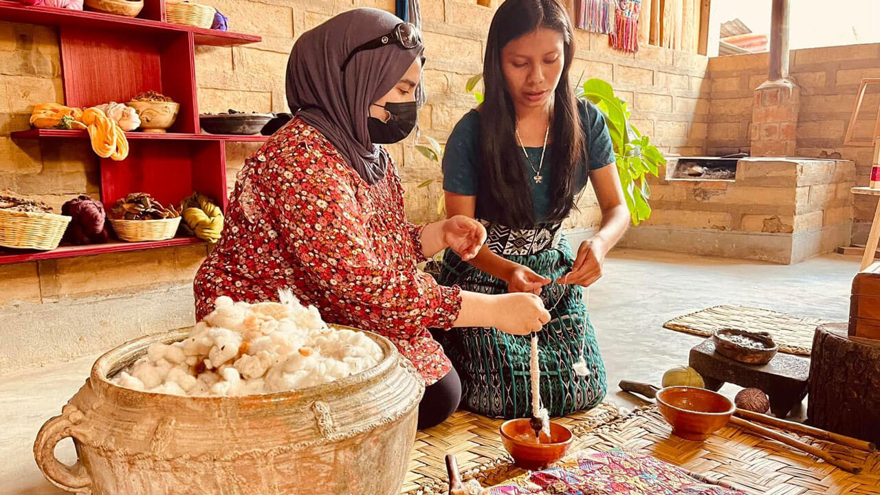 A student kneels on a rug with another woman to learn how to make a cotton thread in Guatemala.