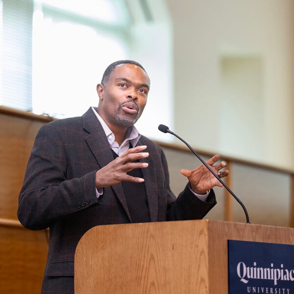 Ron Jackson holds his arms up as he speaks into a microphone at a Quinnipiac podium