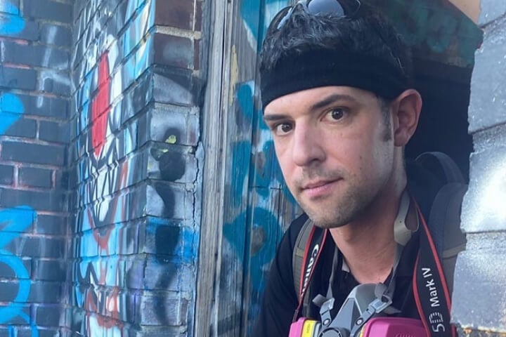 Photo of John Lazzaro leaning on a graffiti-filled wall holding a camera.