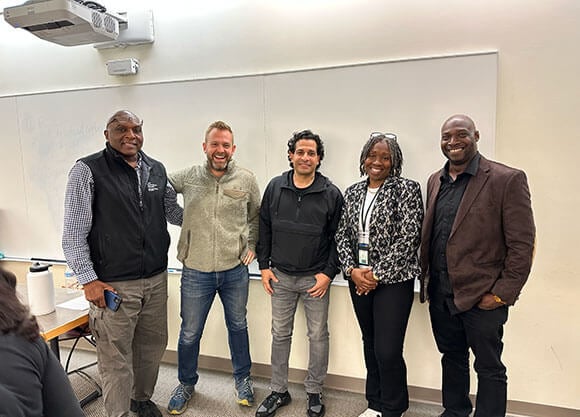 5 individuals smile in front of a white board in a classroom