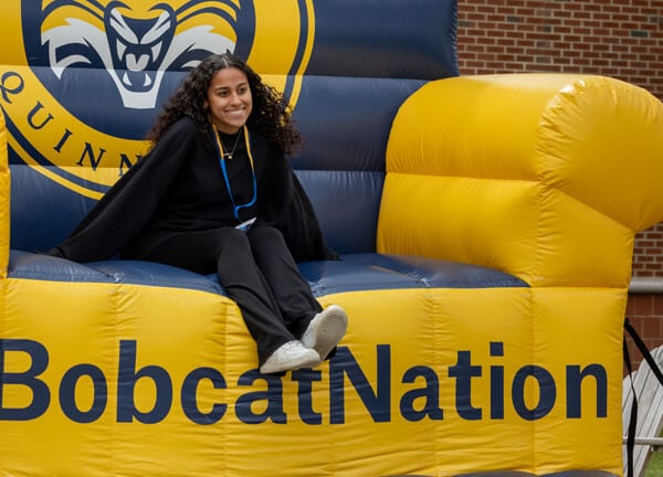 An admitted student sits on a giant inflatable Quinnipiac chair and smiles for a photo