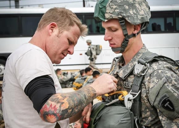 Zach Miller buckles a parachute pack onto a soldier wearing camoflage and a helmet