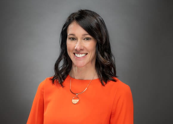 Kori Stewart smiling against a grey background in a vibrant orange blouse