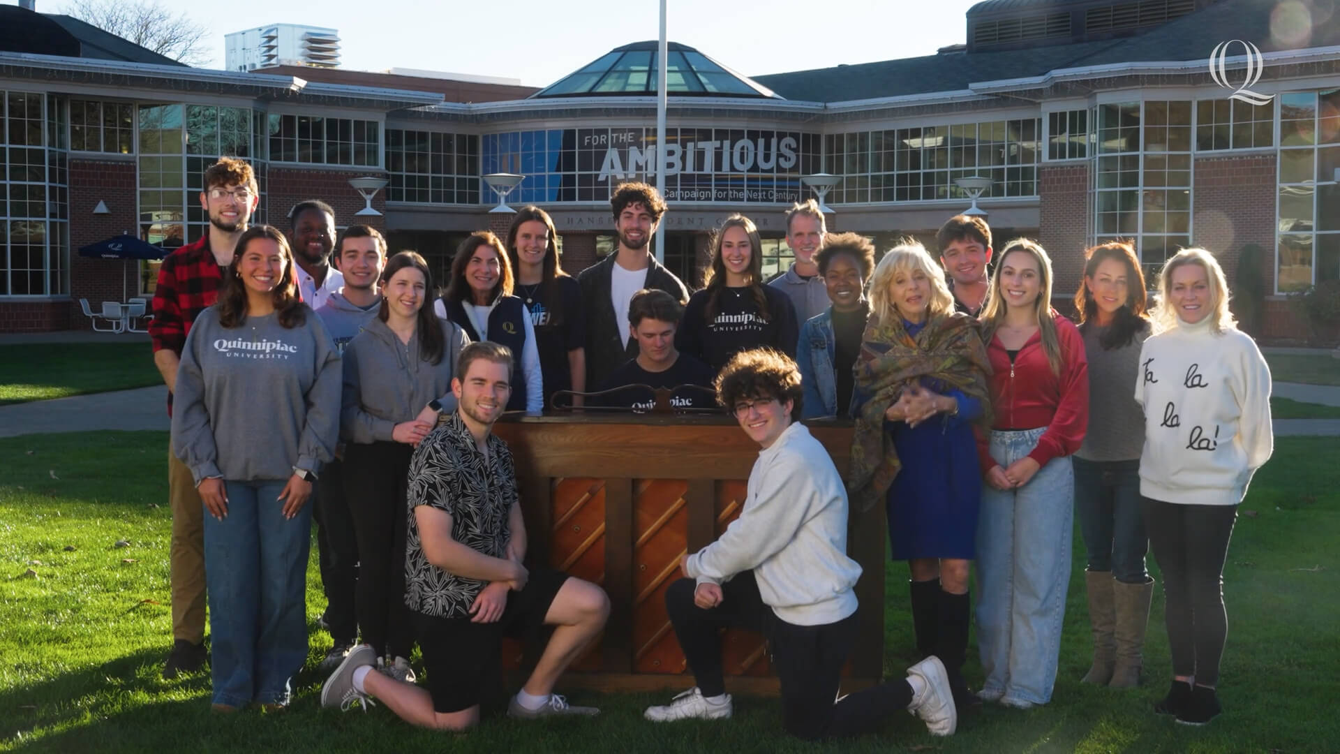 A dozen students and President Olian gather around a piano on the quad