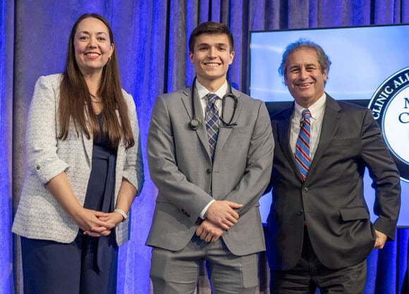 Alex Holle standing in a suit with a stethoscope around his neck, standing next to two other people.