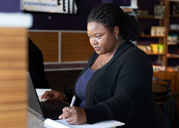 woman writing resume at desk in front of a laptop