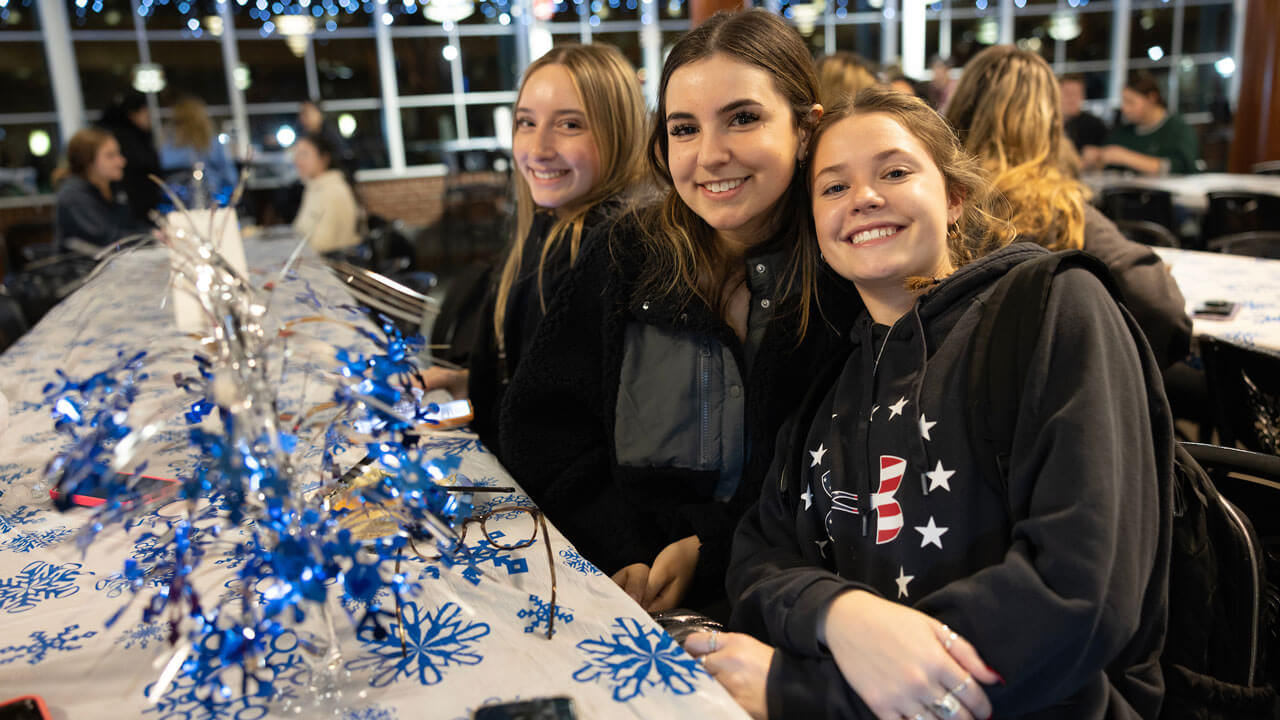 Three students sit together and smile at undergraduate holiday dinner table.