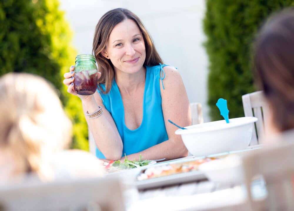 Dana White sitting outside at a table holding a mason jar of juice, looking at two of her daughters