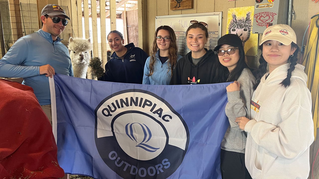 Students hold a flag that says Quinnipiac Outdoors at an Alpaca farm