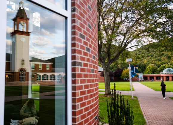 The library clocktower reflects on the student center windows as a student walks across the quad