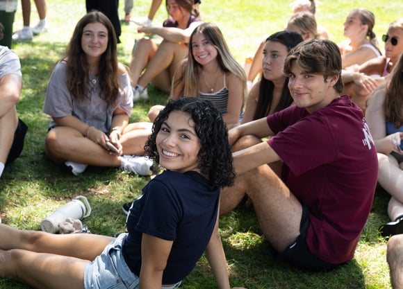 A handful of students sit in a circle in the grass and turn to smile for a photo during the Welcome BBQ