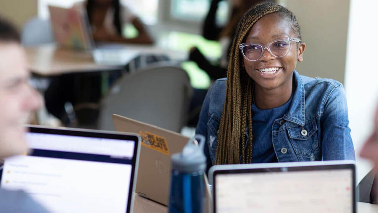 A student smiles while working on her laptop with two classmates at a table in the School of Communications