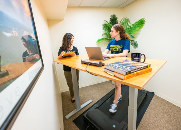 A female student working at a standing desk while on a walking pad being studied by a woman with a clipboard