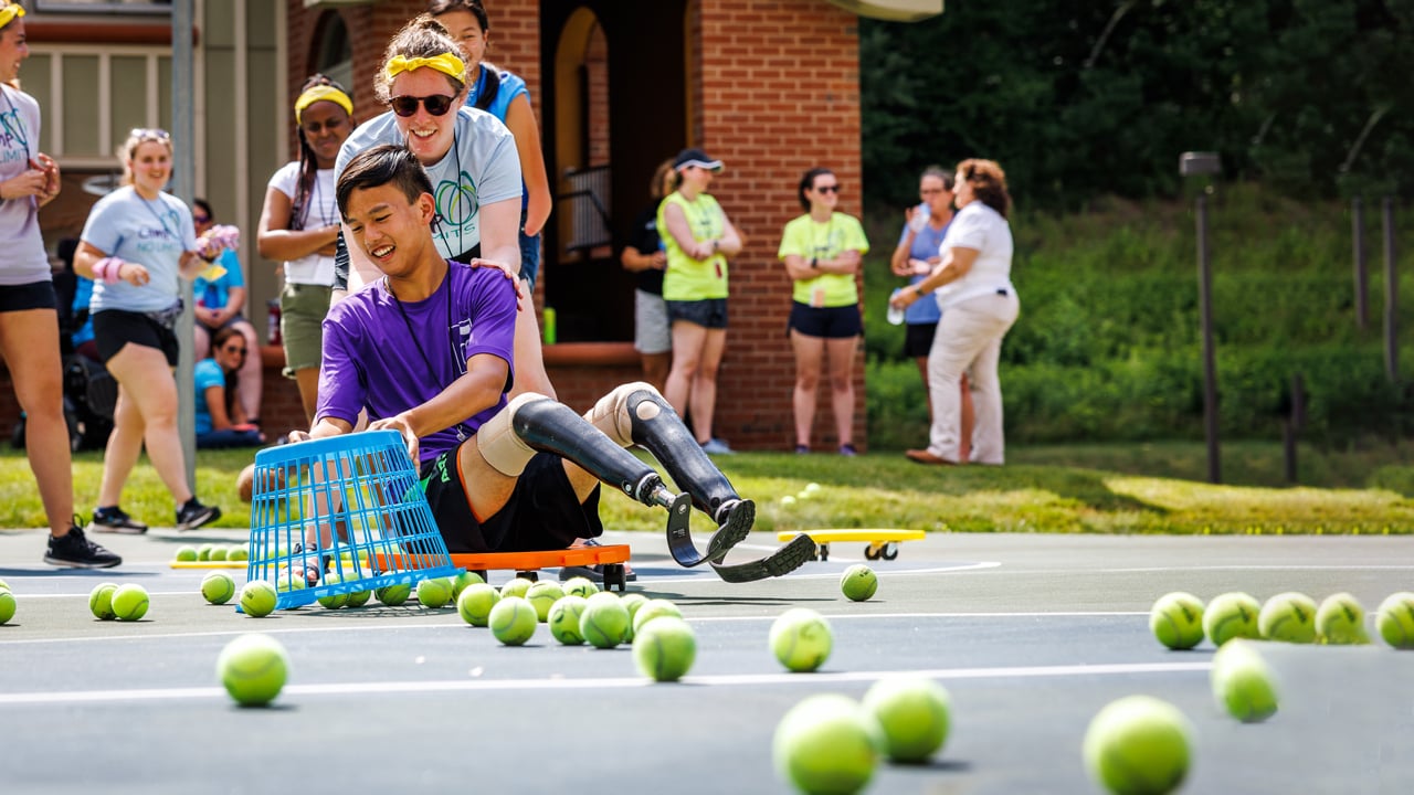 A Camp No Limits volunteer pushes a participate on a roller scooter while the participate collects tennis balls