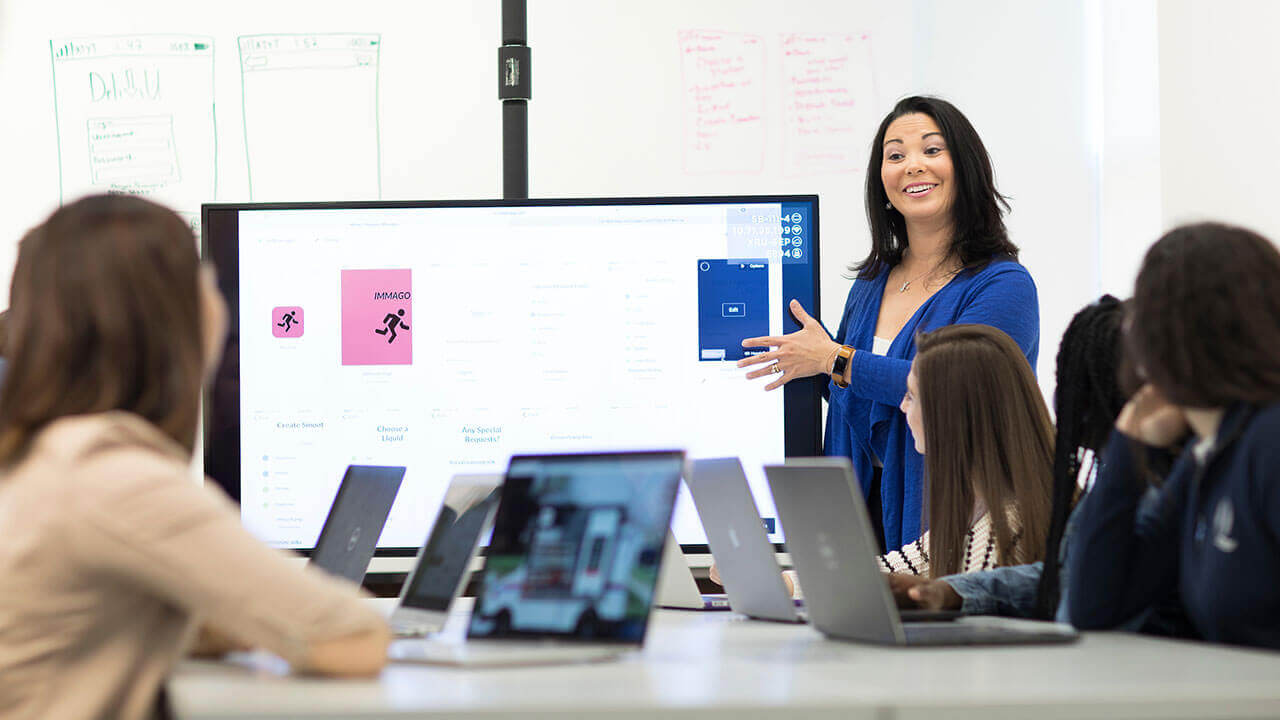 A professor gestures toward a Powerpoint presentation on a large monitor